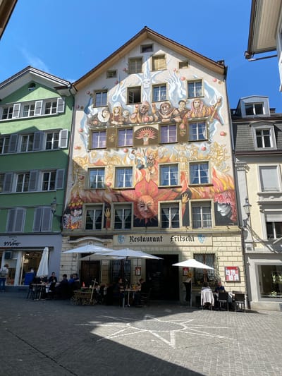 The image depicts the painted facade of Restaurant Fritschia, a building in Lucerne, Switzerland. The building next to it is the Hotel Mimo. The restaurant's exterior is decorated with an elaborate mural featuring figures in historical attire, sun-like faces, and swirling patterns. The building's windows are integrated into the artwork. Patrons are seated at tables under white umbrellas outside the restaurant. The street is paved with cobblestones. Other buildings are seen in the background.