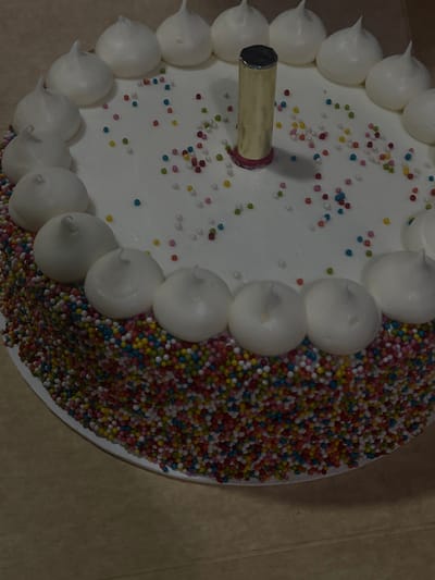 A top-down view shows a round cake with white frosting, adorned with colorful sprinkles scattered across its surface. A ring of white meringue kisses lines the cake's edge. Colorful sprinkles cover the remaining side of the cake. A golden candle is inserted near the center of the cake, completing the celebratory presentation.
