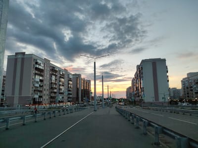 The image captures a wide, paved road in what appears to be a residential area of the city of Pridorozhny, Russia, at dusk. On either side of the road are large, multi-story apartment buildings with many balconies and windows. The sky is filled with dramatic, cloudy formations, with streaks of orange and pink light from the setting sun visible on the horizon, breaking through the darker clouds. Streetlights line the road, some illuminated. Several pedestrian crossing signs are visible. A person on a bicycle is seen on the left side of the road, near a metal guardrail. The overall scene suggests a typical urban environment during the transition from day to night.