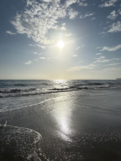 The image shows a beach scene, likely in Cabarete, Dominican Republic. The scene is split horizontally, with the sky and clouds on the left and the ocean on the right. The sun is bright and partially obscured by clouds. The ocean has small waves and reflects sunlight. The beach is not visible. No people are present.