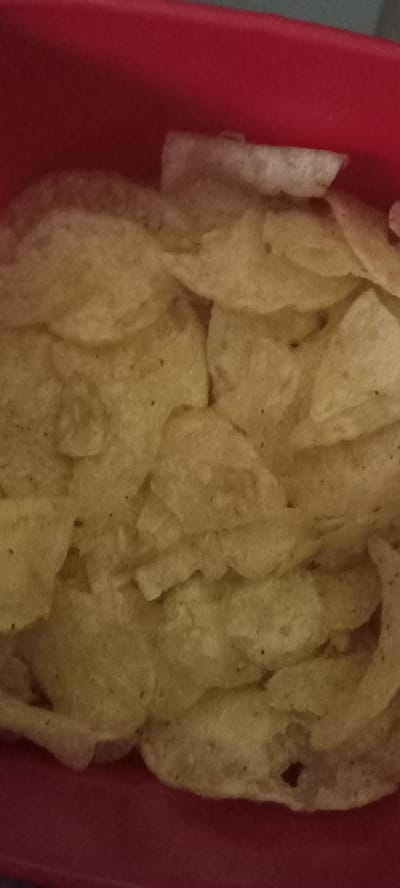 The image shows a close-up view of potato chips in a red container. The chips are golden brown and appear to be slightly greasy. They are piled on top of each other, filling the container. The background is out of focus, but appears to be a dark surface. There are no people or other objects visible in the image. The image does not contain any information about the location in the city of Koviahy, Ukraine.