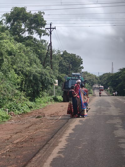 A group of four people are gathered near a utility transformer on a dirt road, adjacent to a paved road in Kothli, India. The three individuals in the foreground, two women and one man, are dressed in traditional Indian attire. The man, wearing a white turban, points towards something off-camera while speaking to the women. The woman on the left wears a floral print dress, and the woman on the right wears a colorful saree. Behind them, a large green utility transformer is mounted on a vehicle. The scene is set against a backdrop of lush green trees and vegetation, with a cloudy sky overhead. In the distance, a bus and several motorcycles are visible on the paved road, indicating ongoing traffic. The dirt road is muddy and has tire tracks, suggesting recent rain or wet conditions. A power line pole with wires is also visible to the left of the scene.