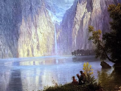 This image captures a large-scale projected or displayed artwork depicting a grand natural landscape, observed within an exhibition space in the city of Reus, Spain. The scene presents towering, rugged mountains rising steeply around a broad, tranquil body of water. A prominent waterfall descends from a high chasm between central mountain peaks, creating a misty effect as it reaches the water below. The mountain faces exhibit varied textures and coloration, illuminated by light that highlights areas in soft yellow, gold, purple, and blue tones.

In the immediate foreground, on a grassy bank at the water's edge to the right, two figures are present. One figure, a male wearing a hat, is seated before an easel with a canvas, engaged in painting or sketching the panoramic landscape. A second, smaller figure is seated slightly behind and to the left of the painter, observing the scene. Sparse bushes and a small tree with distinctive yellow foliage surround the figures, while a larger, darker tree leans over the water further to the right, next to a partially submerged rock. The water's surface reflects the distant mountains and sky, displaying subtle ripples.