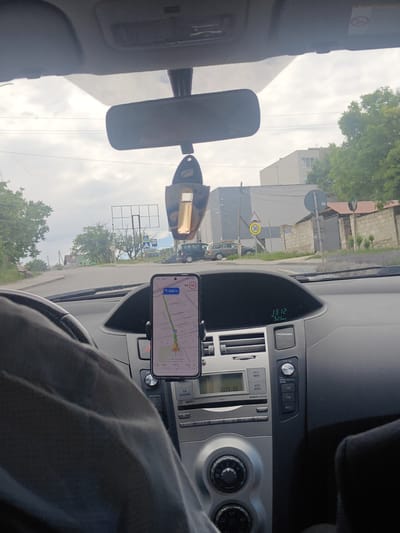 The image is a first-person view from inside a car driving through Colonița, Moldova. A smartphone is mounted on the dashboard, displaying a navigation app with a route and current speed of 50 km/h. The dashboard shows a clock displaying 13:12. The car is moving on a road with buildings and trees visible on either side. A speed limit sign indicating 30 km/h is also present. The sky is overcast. The driver's left arm and shoulder are visible in the foreground.