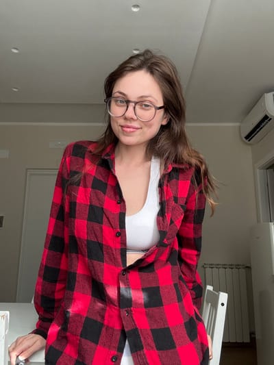 A young adult female is depicted in a residential interior in Scafati, Italy. She stands or leans slightly forward, her left forearm resting on a white table or counter surface in the foreground. Her head is angled slightly to the left, and she looks directly at the camera with a subtle smile. She has long, dark brown hair parted on the right and wears round, clear-framed eyeglasses with dark temples. Her attire consists of an unbuttoned red and black buffalo plaid long-sleeved shirt worn over a white, athletic-style crop top or bralette. A dark band with light text, possibly a brand name, is visible beneath the crop top. The background features light-colored walls. To the left, a portion of a white door frame and a wall-mounted light switch or control unit are visible. To the right, a white split-system air conditioning unit is mounted high on the wall, beneath which a white panel radiator is present. A portion of a white chair back is visible in the lower right, along with a partial view of a white appliance resembling a refrigerator. The white ceiling contains two visible recessed spotlights. Natural light appears to enter from an unseen source, casting shadows within the room.