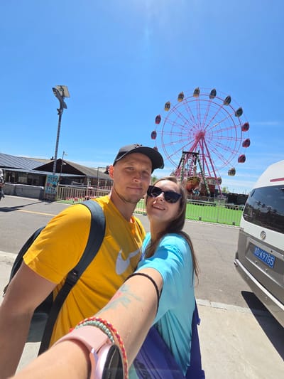 A selfie captures two individuals enjoying a sunny day at an outdoor amusement park or attraction in Jiejinkou, China. The man, partially in the foreground, wears a black cap, a yellow t-shirt with a white logo, and a black backpack. Behind him, a woman smiles brightly, donning sunglasses and a blue t-shirt. She wears a pink smart watch and colorful bracelets on her left wrist, which also shows a tattoo.

The dominant feature in the background is a large, vibrant red Ferris wheel, its structure intricate, holding multiple gondolas that are alternately yellow and dark green/red. To the right, a white van or camper with a Chinese license plate, "冀 D·K97G5", is parked. Behind the couple, buildings resembling food stalls or shops, with some blurry Chinese text on signs, line a paved area. A green fence is visible in the middle ground. The sky is a clear, brilliant blue, indicating bright and sunny weather. A street lamp with a solar panel and a decorative element shaped like a bird or fish is visible on the left. The overall scene suggests a cheerful and leisurely outing.