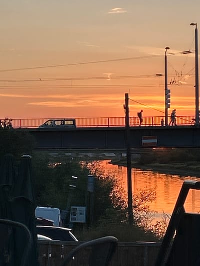 The image depicts a sunset scene over a river in Mannheim, Germany. In the foreground, shadowy figures are seated on chairs, and a white van is parked on the left. A bridge spans the river, with a tram or train visible on it. Several people, including a person with a backpack, are walking on the bridge. The sky is a vibrant orange and yellow, reflecting on the water. Streetlights are visible on the right side of the image.