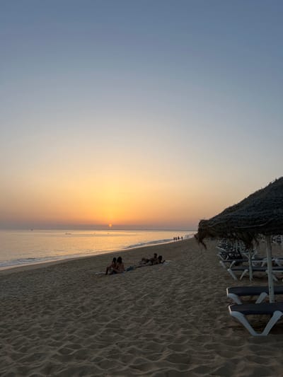 The image displays a beach scene at either sunrise or sunset in Almancil, Portugal. The sky transitions from a light blue at the zenith to a warm orange and yellow gradient near the horizon. The sun's distinct orange disk is positioned just above the horizon line over a calm, reflective ocean. A small vessel is visible in the distant water to the left.

The foreground and mid-ground are comprised of light-colored sand with undulating patterns. Four individuals are present in the mid-ground: two are seated upright on a light-colored mat, facing left; two are lying prone on a mat to their right, facing the ocean. All four appear to be in swimwear. Further along the shoreline, in the distant right, a group of approximately five to seven individuals are walking along the water's edge, appearing as dark silhouettes.

Along the right side of the frame, a continuous line of thatched-roof parasols and associated lounge chairs extends into the distance. The closest parasol is partially visible in the extreme right foreground. The overall scene is illuminated by the soft, warm light of the low sun.
