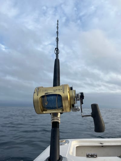 A gold-colored "T-BAHD 50W II S-MAX 70" fishing reel, exhibiting wear with visible scratches, is mounted on a black fishing rod. Blue fishing line is spooled on the reel, marked with a white downward arrow. The rod is secured in a black rod holder on a white fiberglass boat gunwale or transom coaming. A small, hinged white compartment is visible on the boat surface. The scene overlooks dark blue-grey ocean water with visible ripples, extending to an overcast horizon under a sky filled with grey and white clouds. The activity takes place offshore, near Port Kembla, Australia. No individuals are visible.