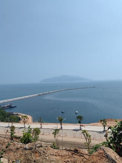 An elevated, wide-angle view captures a coastal scene in Hải Vân Ward, Vietnam. The foreground is composed of reddish-brown earth, rocks, and sparse green vegetation. A newly constructed paved road, bordered by white and red barriers and lined with young trees and streetlights, traverses the scene horizontally below the viewpoint. This road meets a small, light-colored sandy beach and a cove on the left, where a small port facility with two moored industrial vessels is visible. Extending into the calm, dark blue bay water is a long, straight breakwater, constructed from numerous grey rock formations, which stretches from left to right. Two small, light-colored boats are positioned on the open water. In the distant background, a hazy mountain range or island is visible on the horizon, merging with the pale blue sky. Faint, indistinct structures are discernible on the distant coastline to the far right. No individuals are visible.