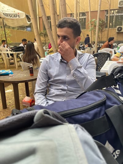 This image depicts an outdoor gathering area in قرية الضبية, Syria. The foreground is dominated by a seated male individual with dark hair and stubble, wearing a light grey long-sleeved collared shirt. He is positioned facing left, with his right hand partially covering his mouth and nose area. In the immediate lower right, a dark blue sports bag with black straps and zippers is prominently visible, partially obscuring the view. A light grey bag is partially visible in the lower left foreground.
The midground features a round brown table to the left of the primary subject, holding a black can with red accents (appearing to be an energy drink) and a blue plastic wrapper. Below this table, a white and red box is on the ground. Multiple other individuals are visible in the background, seated at various white and dark tables with corresponding chairs. One woman with long dark hair is seated with her back to the camera, directly behind the foreground table. Other men are seated further back. The environment consists of dirt ground, several slender tree trunks vertically traversing the scene, and a light-colored building with windows and an external air conditioning unit in the distant background. A white umbrella with "NESCAFE" branding is present on the left side of the scene, partially open.