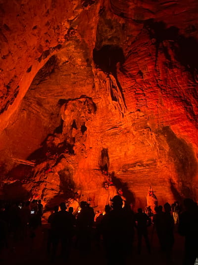 The image depicts a group of people gathered inside a cave, illuminated by vibrant orange lighting that casts dramatic shadows on the rock formations. The cave walls exhibit intricate textures and layered geological structures, with some areas appearing rough and others smoothed by time. In the background, at the center of the cave, is a large rock formation resembling a sculpture or a deity, with smaller figures or deities positioned around it. The people are silhouetted against the bright light, suggesting they are observing the illuminated features within the cave. The environment is dark and cavernous, characteristic of a natural cave system. The location is identified as the cave in Gollagutta, India, a place known for its religious significance and natural beauty, often featuring such illuminated rock formations and spiritual iconography.