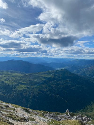 The image depicts a panoramic view of a mountainous landscape, likely captured from an elevated viewpoint. The foreground consists of a rocky, sparsely vegetated slope with a winding path. In the middle ground, rolling green hills covered in dense forest dominate the scene, with several small structures or dwellings visible. Further in the distance, successive layers of mountains fade into a hazy blue, suggesting significant depth and atmospheric perspective. The sky is partly cloudy, with a mix of white cumulus clouds and some darker, more ominous formations against a bright blue sky.

While the exact location is not visually verifiable from the image alone, the description suggests it is a rural mountainous area. Given the provided location of Katun Vulića, Montenegro, the scene is consistent with the mountainous terrain of that region. There are no identifiable people or specific actions depicted in the image. The focus is on the natural environment and its vast scale.