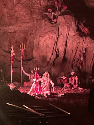 The image depicts a scene within a cave in Gollagutta, India, likely at the Birla Mandir complex. In the foreground, a dark, possibly wooden structure with horizontal beams is visible. The midground features several figures, appearing to be mannequins or statues, in a cavernous setting illuminated by red lighting. Two figures are prominent in the center, one in a red garment and another with long, white hair. Both are positioned in a seated or kneeling posture. Several tridents, religious symbols associated with Hinduism, are placed throughout the scene, with one being held by a figure. In the background, more figures are situated on ledges and higher ground within the cave. The environment is rocky and uneven, characteristic of a natural cave formation.