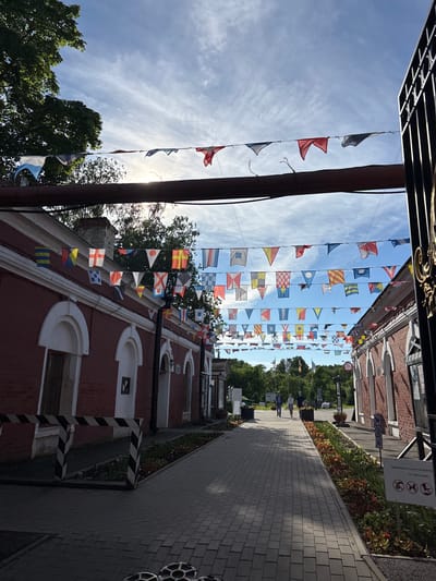 The image depicts a street lined with red brick buildings, likely in Kronstadt, Russia, given the navigational flags adorning the street and the style of architecture. The buildings feature white arched window frames and decorative cornices. A paved walkway runs between the buildings, bordered by flowerbeds. Several individuals are walking along the path in the distance. The sky is bright blue with scattered clouds, and sunlight filters through the numerous navigational flags strung across the street, creating a festive atmosphere. To the right, a black metal gate or fence with ornate details is visible. Signs are posted near the buildings, one with Russian text and symbols indicating rules for visitors. The overall scene suggests a well-maintained public area, possibly a tourist destination or a historic district.