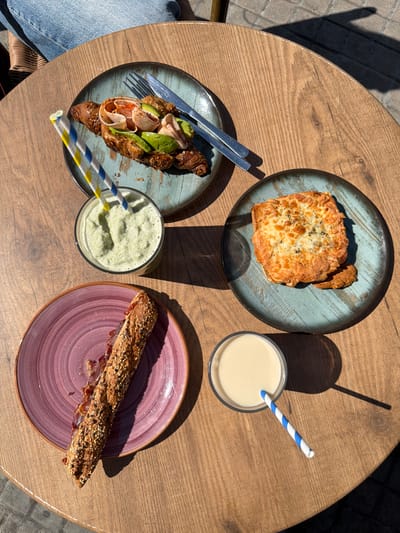 The image shows a round, wooden table with four plates of food and two glasses of drinks, likely taken in an outdoor café or restaurant. On the top left is a blue plate holding a square piece of cheesy bread, next to a glass with a white liquid and a blue and white striped straw. The bottom left contains a blue plate holding a croissant topped with avocado slices and thinly sliced meat, along with a fork and knife, next to a green drink with a yellow and blue striped straw. The bottom right plate, colored in shades of pink, holds a long, narrow pastry covered in seeds. The scene is brightly lit, indicating a sunny day. The image captures a moment of enjoying a meal. It is unlikely to be in the city of Burjassot, Spain.