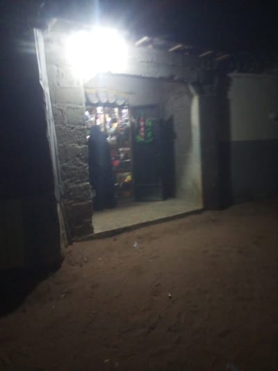 The image depicts a dimly lit scene at night, possibly in a rural or less developed area of Yola, Nigeria. In the center of the frame, a small shop or market stall is visible, illuminated by a bright overhead light. Shelves stocked with various goods, appearing to be packaged items and possibly drinks, are discernible behind a counter. A person, silhouetted in dark clothing and wearing a headscarf, stands behind the counter, suggesting they are the shopkeeper or a customer. The entrance to the shop is framed by rough, possibly brick or concrete, walls. The ground in front of the stall is composed of loose soil or sand, with some scattered debris. To the left, a darker, more solid structure is present, partially obscuring the view of the shop's interior. The overall impression is of a humble, functional establishment operating in the evening hours.