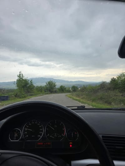 The image provides a driver's-seat perspective from inside a car, looking out through a rain-speckled windshield at a rural landscape. A winding asphalt road is visible through the windshield, extending into a valley surrounded by green, tree-covered hills and distant, hazy mountains under an overcast sky. Numerous raindrops are visible on the windshield, suggesting recent or ongoing precipitation.
The right side of the image features a portion of the car's interior, including the steering wheel and dashboard. The speedometer on the dashboard registers 0 km/h, indicating the vehicle is stationary or moving very slowly. The tachometer shows the engine idling at approximately 500 RPM (0.5 x1000 1/min). The odometer displays a total mileage of "288282 km", with a trip meter reading of "279.9 km". The fuel gauge indicates the tank is slightly above empty, and the word "DIESEL" confirms the fuel type. Several warning lights are illuminated on the dashboard, including a red general warning symbol (exclamation mark in a circle), an orange light that resembles an engine or system fault indicator, and another red light, possibly for brakes or another critical system.
The steering wheel, with a textured grip, is visible in the foreground. The weather is cloudy and appears to be daytime, with a subdued light cast over the mountainous or hilly terrain. The scene depicts a quiet, scenic road in a rural setting, consistent with the given location context of Lelintsi, Bulgaria.