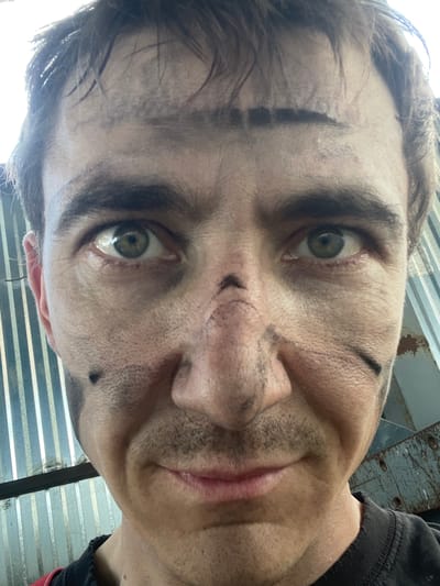 The image is a close-up, low-angle, upside-down selfie of a person's face. The subject appears to be a male with light skin, green eyes, and dark hair. His face is smudged with dirt or grease, particularly around his eyebrows, nose, and cheeks, suggesting he has been engaged in some kind of manual labor or outdoor activity. There are dark smudges that resemble war paint or decorative markings on his forehead, nose, and chin. He is smiling slightly, with his mouth open, and his eyes are wide and looking directly at the camera, conveying a sense of excitement or mischievousness.
The background is out of focus but appears to be an industrial or outdoor setting. There are corrugated metal sheets visible, suggesting a wall or structure. The lighting indicates it is daytime, possibly bright sunlight filtering in, as suggested by the bright illumination on the left side of the subject's forehead. The overall mood is informal and energetic. The provided location context of Сокуры, Russia, suggests the image was taken there. There is no visible text in the image.