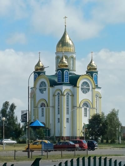 The image depicts the Resurrection Cathedral in Kobryn, Belarus, on a clear day with scattered clouds. The church, a prominent architectural landmark, features a central golden dome topped with a cross, flanked by smaller golden domes. The building's facade is a mix of white and pale yellow vertical stripes, with arched windows adorned with blue panes. A green roof caps the main structure, with additional green accents.

In the foreground, a black metal fence separates a street from a grassy area. Several vehicles are visible: a yellow taxi, a red hatchback, and a dark-colored SUV parked in front of the church. A silver sedan is parked to the left. Trees line the background, with a tall lamppost and a signpost to the left of the church. A bright blue canopy is visible near the entrance of the church. The overall scene suggests a typical day in the city, with religious and everyday elements coexisting.