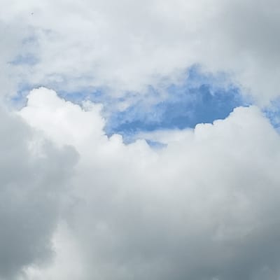 The image shows a section of the sky, predominantly filled with white and gray clouds. A patch of light blue sky is visible through a break in the cloud cover. There are no people, animals, or objects. The location is unidentifiable from the image content. There are no identifiable actions or interactions. The environment is cloudy.