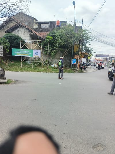 A street view shows a man in a safety vest standing at an intersection. There are several vehicles visible further down the street. A building with a tiled roof sits on the left side of the image, and a banner is propped up on a bamboo frame in front of it. Wires crisscross the sky, and a blurry object is visible in the foreground.