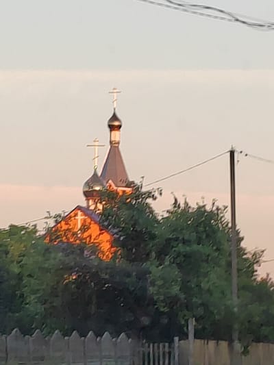 A soft focus, low-resolution image depicts the upper portion of an Orthodox church structure peeking through a dense canopy of green trees. The church features a tall, dark gray, conical roof topped with a golden dome, and a smaller, rounded dome also topped with a golden dome. Two ornate crosses, one on the main dome and one on the smaller dome, are visible. A third, smaller cross with a white outline is affixed to the reddish-orange facade of the lower part of the church, which is partially obscured by the foliage. The sky is a soft, pale orange, suggesting either dawn or dusk. In the mid-ground, a dark wooden utility pole with wires running across the frame is present. A blurred, light gray wooden fence runs along the bottom of the image. The image is shot from a low angle, looking up at the church.