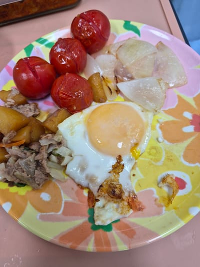 This is a top-down, close-up, medium shot of a colorful, floral-patterned plate filled with food. The plate contains four cherry tomatoes, which appear to be roasted or stewed. There are also pieces of what look like fried onions or leeks. In the center of the plate is a sunny-side-up fried egg with a runny yolk. To the left of the egg is a portion of what appears to be shredded meat mixed with diced potatoes and possibly some cabbage or other vegetables. The food is arranged on a pink table. The overall impression is of a home-cooked meal. No specific location in Suwon-si, South Korea can be determined from the image.