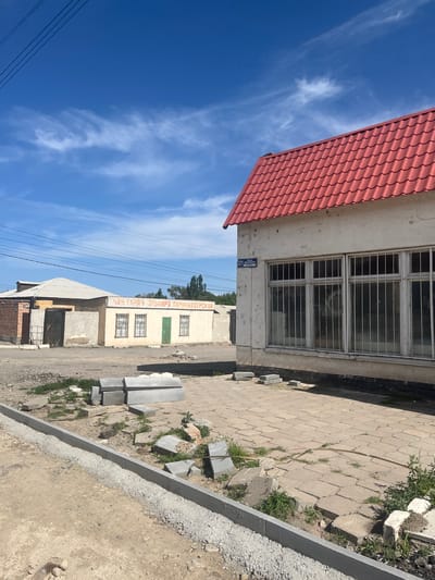 The image shows a street scene in Balykchy, Kyrgyzstan. In the foreground, a newly constructed concrete curb is visible, with loose gravel and some broken concrete pieces nearby. Beyond the curb, a paved area with interlocking hexagonal stones is present, overgrown with grass in some sections. Stacked concrete blocks and some scattered construction debris are also scattered on the paved area.
To the right, a single-story building with a white facade and a red tiled roof is prominent. The building has large windows covered with metal bars. A blue street sign with white text is affixed to the left side of this building. Further to the left, another building with a lighter colored facade and a flat, light-colored roof is visible, with a brick structure attached to its left side. This building has multiple windows and a large green metal gate. Red Cyrillic lettering is visible across the facade of this second building, appearing to be a sign for a business. The sky above is blue with some wispy white clouds, and electrical wires are strung across the upper portion of the image. The overall scene suggests ongoing construction or maintenance work in a semi-urban or rural area.
