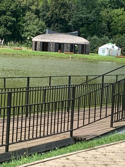 The image shows a calm body of water with a wooden walkway and black metal railings in the foreground. In the background, there is a long, dark-colored building with a curved roof and several open doorways. To the right of this building is a white tent with green accents and a green emblem, resembling a yurt. A person can be seen near the tent. The area is surrounded by lush green trees. The location is identified as Arbuzov-Baran, Russia.