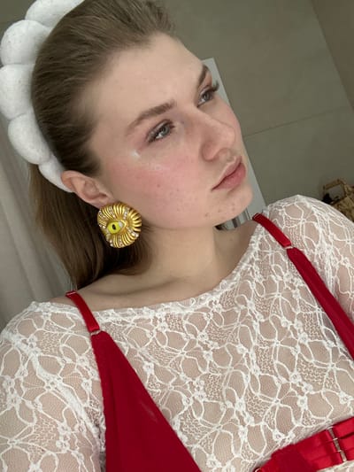 A woman with light skin and brown hair poses. She wears a white lace top with red straps. A large, gold, eye-shaped earring adorns her ear. Her head rests on a white, floral-patterned cushion or headband. The background appears to be a plain, light-colored surface.