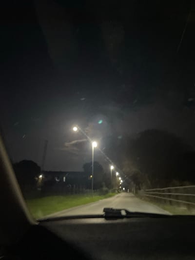 The image captures a nighttime scene from inside a vehicle, looking forward through the windshield, revealing the dashboard and a windshield wiper in the lower foreground. The setting is a wet, paved road in Monsummano Terme, Italy, extending into the distance and illuminated by a series of bright streetlights positioned along its left edge. To the left of the road, a grassy verge precedes a fence, beyond which faint outlines of a low-rise building and a tall, slender industrial structure are visible against the dark sky. The right side of the road features a wooden fence running parallel to the asphalt, backed by dense foliage or trees. The night sky is dark, with scattered clouds partially obscuring a prominent, bright light source, likely the moon. No individuals or specific actions are discernible beyond the implicit presence of the vehicle's occupant.