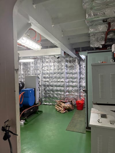 The image depicts the interior of what appears to be a mechanical room or engine room, possibly on a vessel or industrial facility, with a green floor. Prominently featured is a blue and black generator on a green base. The walls and ceiling are covered with silver, quilted insulation material. In the background, there is a large, light green electrical panel. To the right of the generator, there is a red bucket containing orange rope, and next to it, a pile of wood and rope. A fluorescent light fixture is visible on the ceiling. The image is a photograph taken from a medium distance, showing the room's contents and construction. The location in Melaya, Indonesia, is not visually identifiable within the image itself, as there are no distinguishing landmarks or elements that would indicate this specific geographical location.