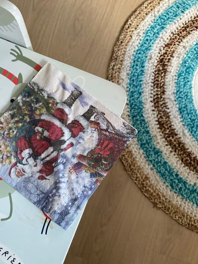 A close-up, high-angle shot shows a festive napkin with a Christmas-themed print featuring multiple Santas and snow-covered trees. The napkin is placed on a light blue surface with a cartoon character's arm and a red and green striped sleeve visible on the left. To the right of the napkin is a circular, crocheted rug with alternating stripes of white, light blue, and brown. The rug is positioned on a wooden floor. The scene appears to be indoors, likely a home environment. There is no identifiable information to link this image to Osek, Czechia.