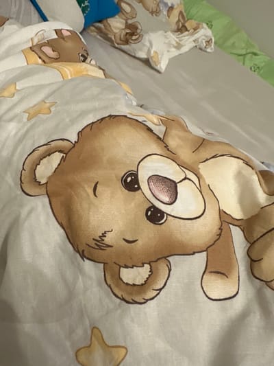 The image shows a close-up of a duvet cover with a cartoon teddy bear print. The duvet cover is light-colored with a repeating pattern of teddy bears and yellow stars. The main teddy bear visible in the foreground is brown with large, dark eyes and a pink inner ear. The background appears to be a bed with a gray sheet. The context of Malinovka, Russia, is not discernible from the visual information provided in the image.