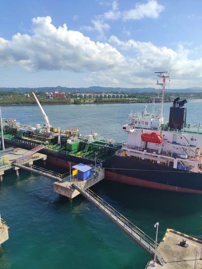 This elevated view captures a large tanker or cargo ship docked at a pier in Kunak, Malaysia, under a bright daytime sky with scattered white clouds. The vessel, featuring a black hull, a red boot-topping line, and a white superstructure, dominates the right side of the image. Its deck is equipped with numerous green and yellow pipes, tanks, and machinery, including a long white crane extending over the water.

A metal gangway with yellow railings provides access from the ship to the concrete pier. The pier itself has yellow safety markings and multiple bollards for mooring. The water in the harbor is a deep teal-green, appearing calm with some subtle ripples. In the distance, along the opposite shore, a long stretch of industrial facilities and buildings with cylindrical storage tanks can be seen, backed by green hills or mountains under a hazy sky.

A red enclosed liferaft is prominently visible on the ship's superstructure, marked with a white cross. Other details on the ship include various antennae and masts. The overall scene suggests a routine port operation, likely involving the loading or unloading of cargo from the tanker. No people are discernible in the image. Visible text is limited; a white cross is on the liferaft, and faint, illegible text appears on small signs on the ship's deck and superstructure. The bright sunlight indicates clear and fair weather conditions.