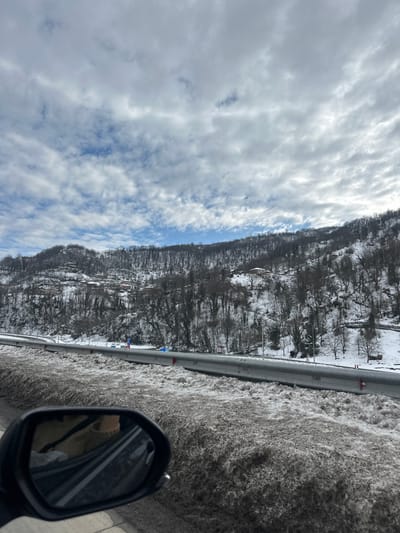An outdoor, eye-level photograph captured from within a vehicle shows a snow-covered mountainous landscape in Ubisa, Georgia, under a partially cloudy sky. In the foreground, the side mirror of the vehicle is visible on the left, reflecting a partial view of an occupant wearing light-colored clothing. Adjacent to the vehicle and running parallel to the road is a snowbank composed of dirty, slushy snow. A grey metal guardrail separates this snowbank from a multi-lane highway, where blue and red markers are visible along the shoulder. Beyond the highway, a steep mountainside rises, densely covered with leafless deciduous trees and patches of snow. Several residential structures with dark roofs are scattered across the snow-dusted mountainside. The sky above is largely covered by stratocumulus clouds, with intermittent patches of clear blue sky visible. The overall scene suggests a winter day with recent snowfall.