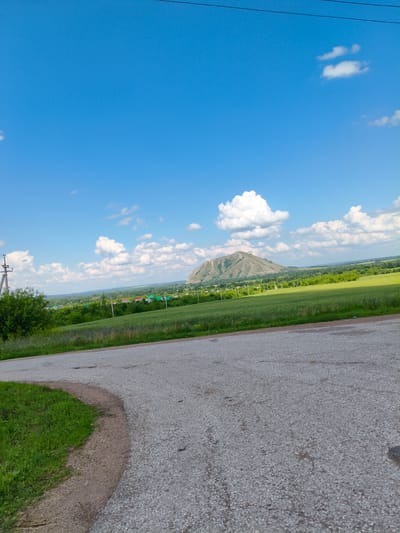 The image depicts a rural landscape under a bright blue sky with scattered white clouds. In the foreground, a paved road curves to the left, with a strip of green grass and soil on its edge. The road appears to be in fair condition with some visible wear and tear. Beyond the road, a vast green field stretches towards a distant village. The village is characterized by scattered houses with colorful roofs, nestled amongst trees. In the far distance, a prominent, conical hill dominates the horizon, its slopes covered in vegetation. Power lines are visible across the top of the image, and a utility pole stands on the left side. The scene conveys a sense of tranquility and natural beauty, consistent with a location in the countryside near Bel'skoe, Russia.