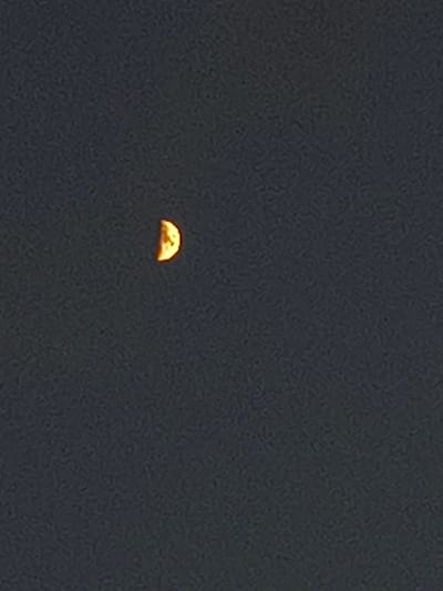 A small, crescent-shaped object, colored orange and yellow, is visible in the upper left quadrant of a dark, textured background. The object appears to be the moon.