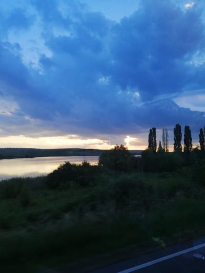 The image depicts a landscape scene at dusk. A large body of water, likely a lake or river, occupies the middle ground, reflecting the muted light of the setting sun. The horizon is a band of pale yellow and orange, transitioning into a dark blue sky filled with dramatic, heavy clouds. In the foreground, a dark, blurred line of vegetation and trees runs across the bottom of the frame, suggesting movement, possibly from a vehicle. The overall impression is one of a tranquil, overcast evening. The location is Pocoleni, Romania.