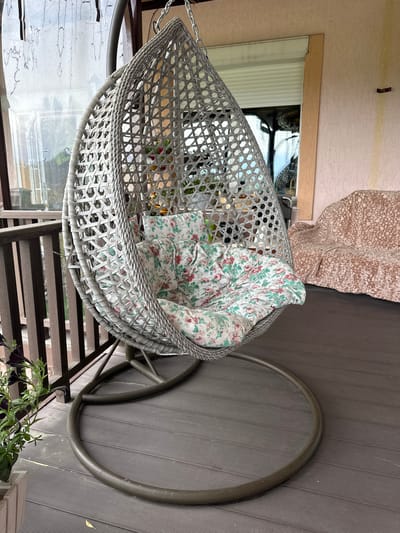 An outdoor hanging chair with a floral cushion sits on a dark wood deck. The chair is made of grey woven material and is suspended by chains from an unseen support. To the right of the chair, a light-pink sofa is covered with a textured throw blanket. In the background, a building with light-colored siding and a roll-down security shutter is visible. A wooden railing is present to the left of the chair. A potted plant with dark purple flowers is on the deck in the lower-left corner of the image. The scene is likely located on a porch or balcony in the city of Bolshaya Rechka, Russia.