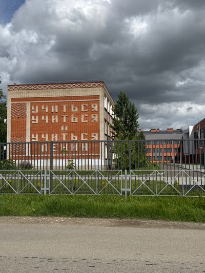 The image depicts a building in Yelizavetinskaya, Russia, near a fenced walkway. The building has a brick facade, and the words "АЛЕКСАНДРОВСКАЯ СЛОБОДА" are written vertically on the building. The sky is cloudy and overcast. The fence is made of metal. A paved path is visible next to the fence. A strip of grass grows between the fence and the path. In the background, another building is visible. A tree is between the building and the fence.