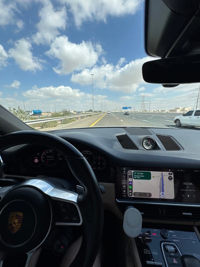 The image displays a driver's perspective from inside a Porsche vehicle on a multi-lane highway in Al Aweer, United Arab Emirates. The time is approximately 13:18 under a bright blue sky with scattered white cumulus clouds. In the foreground, the black multi-function steering wheel, bearing the distinctive Porsche crest, is prominently visible. The vehicle's dashboard features a central infotainment screen operating Apple CarPlay, which shows a navigation map indicating a route to "Sharjah - Kalba M E102" with 5.7 kilometers remaining, and the vehicle's current speed at 102 km/h. A circular chronometer is mounted on the dashboard above the climate control panel, which displays a temperature setting of 22.5°C. A white circular magnetic phone mount is attached to an air vent below the infotainment screen.
Through the windshield, the paved highway extends into the distance, bordered by guardrails and numerous light poles. Other vehicles are visible on the road, including a white pickup truck on the far right lane and a blue tanker truck parked on the left shoulder. The surrounding environment is characterized by an arid landscape with sparse low-lying vegetation. Large electrical transmission towers are visible in the distant background on the right side of the highway.