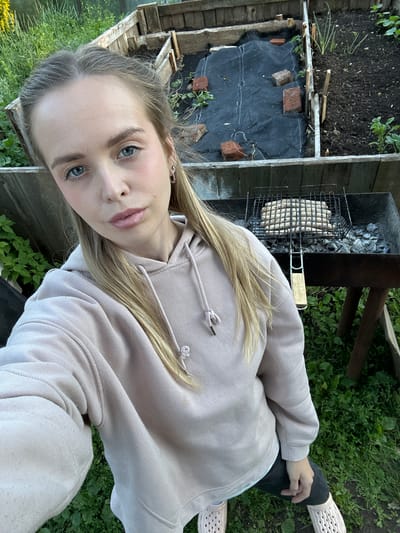 A young woman with blonde hair and blue eyes is taking a selfie outdoors. She is wearing a light pink hooded sweatshirt and dark pants, with light-colored perforated shoes. She is standing in what appears to be a backyard or garden area, with raised garden beds and greenery visible in the background. A barbecue grill with sausages on it is positioned in front of her. The environment suggests a rural or suburban setting. The location is identified as Azinsky, Russia.