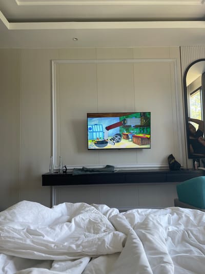 This image captures an interior scene, likely a hotel room or bedroom, in Kayuambon, Indonesia, viewed from a low perspective on a bed. The foreground consists of rumpled white bedding and a white duvet, occupying the lower third of the frame. Above the bed, a dark-colored floating console with multiple drawers is mounted against a light beige paneled wall featuring decorative white molding. On the console, from left to right, are two empty clear glass bottles, a pair of dark-framed eyeglasses, a folded garment resembling dark green or blue fabric, and a dark bag or pouch.

Centered on the wall above the console is a flat-screen television displaying an animated scene, specifically from SpongeBob SquarePants, showing a contraption with pipes, a large red arm, and various objects in a laboratory-like setting. To the right of the television, a tall, arched mirror is mounted vertically. Reflected in the mirror is a person, presumably the photographer, with dark hair and wearing a dark top, looking into the mirror. The reflection also reveals bright sunlight and green foliage outside a window, suggesting a natural outdoor view. The walls are uniformly paneled in a light beige tone with vertical and horizontal white molding accents. The ceiling features a white tray design with a single recessed light fixture visible in the upper center. A portion of a teal or turquoise upholstered chair or ottoman is visible at the far right edge of the frame.