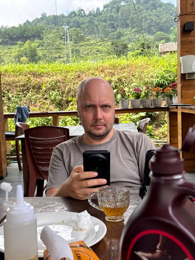 A bald man with a short beard and a neutral expression looks directly at the camera while holding a black smartphone with a prominent circular camera module in his right hand. He is seated at a wooden table in an outdoor or semi-outdoor setting, likely a balcony or terrace, against a backdrop of lush green mountainous terrain, characteristic of Du Già Commune, Vietnam.

On the table in front of him, there is a clear glass mug filled with an amber liquid, possibly tea or beer. To the right, a white plate holds remnants of a meal and a crumpled white napkin. A small white squeeze bottle, likely containing a condiment, sits on the table, along with an orange and white packet partially visible, bearing the text "LAMO". A large, dark brown bottle with a maroon cap featuring a red ring is prominently placed in the foreground, suggesting a topping like chocolate syrup.

The background reveals dense, vibrant green foliage and distant mountains under what appears to be an overcast sky, indicating daytime. Wooden beams and structures frame the scene, and a brown chair is partially visible behind the man. The overall impression is a casual meal or break in a serene, natural environment.