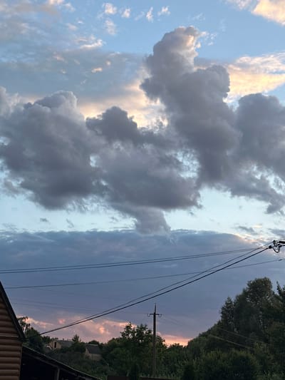 This is a photograph taken in Zvenyhorodka, Ukraine, likely during the late afternoon or early evening, as suggested by the soft, diffused light and the colors in the sky. The image is oriented vertically, capturing a dramatic sky filled with a mix of dark, heavy clouds and lighter, wispy ones. Some of the clouds are tinged with shades of yellow and pink, hinting at the setting sun.
The scene includes elements of a rural or semi-rural setting. Power lines stretch diagonally across the frame, supported by utility poles, indicating the presence of infrastructure. In the upper right and along the right edge of the image, dense green trees form a natural border, with glimpses of rooftops and structures suggesting a village or settlement nestled amongst the foliage. The bottom part of the image shows the edge of a wooden structure, possibly a roof or building, adding to the sense of place.
There are no people visible in the photograph. The focus is primarily on the atmospheric conditions of the sky. The weather appears to be changing, with the contrast between the dark and light clouds creating a dynamic and somewhat dramatic visual. The overall mood is one of natural beauty and quietude, with a hint of the approaching evening. No text is visible in the image.