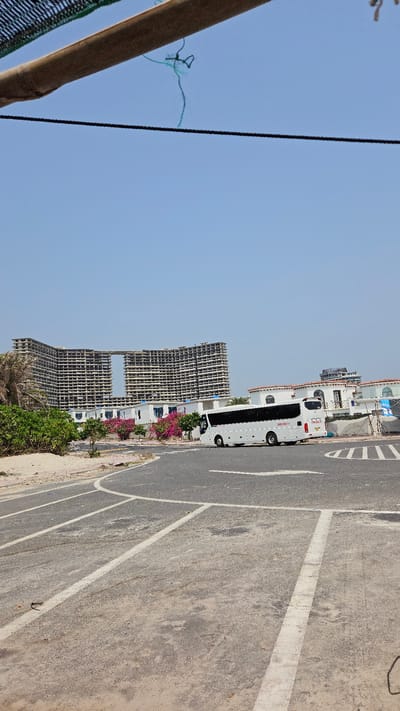 The image captures a bright, sunny daytime scene in what appears to be a developing urban or coastal area, consistent with the context of Bac Cam Ranh, Vietnam. The photo is rotated, presenting the horizon vertically.

In the mid-ground, a white double-decker bus is parked or stopped on the side of a paved road. On the side of the bus, red text reads "FUN&SUN.RU" and black numbers "200-30-77" are visible. In the background, several large, multi-story buildings are under construction, appearing as concrete skeletal structures with many open windows, indicating an ongoing development project. Adjacent to the road and in front of the construction, there are smaller, completed white buildings with red-tiled roofs, surrounded by vibrant pink flowering plants and green bushes.

The foreground shows a paved road with clear white painted lines, possibly marking parking spaces or traffic lanes, alongside a dirt or sand shoulder. To the far left, a wooden post or beam is partially visible, with a dark cable extending upwards (horizontally in the rotated image) and some tangled green string against the expansive, clear blue sky. No people are visible in the scene. The bright, clear sky suggests fair weather.
