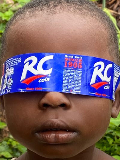 The image is a close-up, eye-level shot of a young child's face, centered vertically and horizontally. The child's eyes are covered by a blue and white paper band, which appears to be a label from an RC Cola product. The band has the "RC cola" logo prominently displayed, along with text such as "Great Taste Since 1905" and "Royal Crown". The child's dark skin tone and the texture of their hair are clearly visible. The background is softly blurred and consists of green foliage, suggesting an outdoor setting, possibly in a rural or natural environment. The image composition focuses tightly on the child's face, drawing attention to the unusual placement of the RC Cola band over their eyes. There is no specific indication of the city of Ufu Esioni, Nigeria within the image itself.
