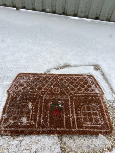 The image depicts an outdoor scene in Zeda Sakara, Georgia, under snowy conditions. In the foreground, a rectangular doormat, brown in color and textured, is shaped and patterned to resemble a gingerbread house. The mat features light-colored outlines for roof tiles, windows, a door, and a small red and green wreath design centrally located on the door area. Light snow accumulation is visible on the surface of the doormat. Surrounding the doormat and extending into the midground is a layer of fresh, undisturbed white snow covering what appears to be a paved or tiled ground surface. In the background, a light-colored, possibly corrugated metal or panel wall or fence is visible, also partially covered with snow or frost.